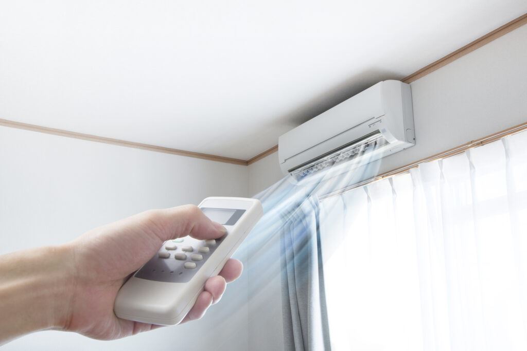 Hand pointing remote at ductless mini-split system on a wall above a window.